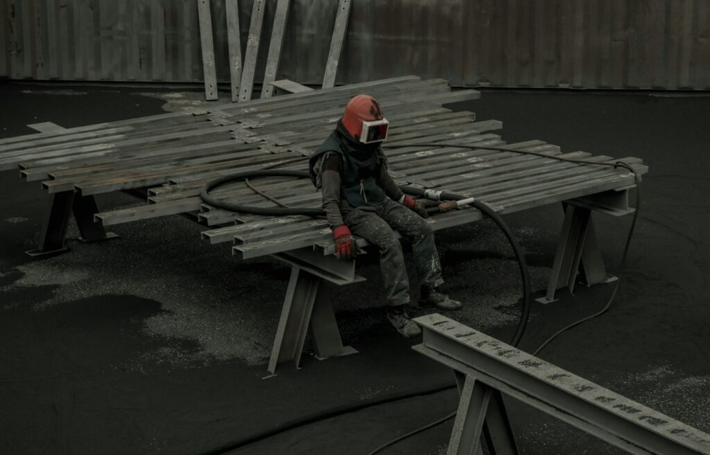 Abrasive blaster in a protective hood sitting on a rack of blasted steel sections